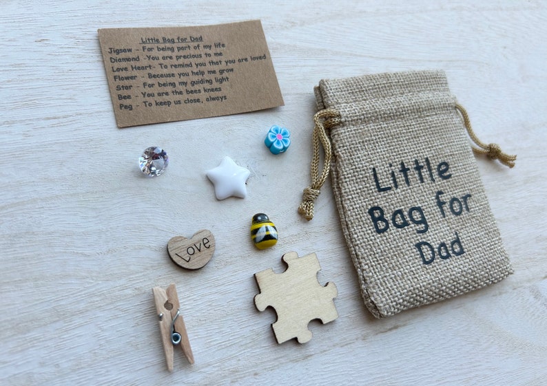 May include: A small hessian bag with the text "Little Bag for Dad" printed on it. The bag is filled with small trinkets, including a wooden heart with the word "Love" on it, a wooden jigsaw puzzle piece, a wooden peg, a white star, a blue flower, a clear crystal, and a yellow and black bee.