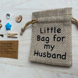 May include: A small burlap bag with a drawstring closure. The bag is decorated with the text "Little Bag for my Husband". The bag is surrounded by small decorative items, including a wooden puzzle piece, a blue star, a small wooden heart with the word "Love" on it, a small crystal, a key, and a clothespin.