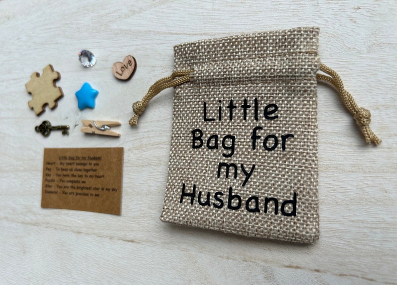 May include: A small burlap bag with a drawstring closure. The bag is decorated with the text "Little Bag for my Husband". The bag is surrounded by small decorative items, including a wooden puzzle piece, a blue star, a small wooden heart with the word "Love" on it, a small crystal, a key, and a clothespin.