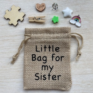 Może przedstawiać: Mały jutowy woreczek ze sznurkiem do zamykania. Woreczek jest ozdobiony napisem "Little Bag for my Sister". Woreczek jest otoczony r&oacute;żnymi małymi przedmiotami, w tym drewnianym sercem z wygrawerowanym napisem "Love", małą zieloną koniczyną, białą gwiazdą, zawieszką w kształcie tęczy i drewnianym elementem układanki.