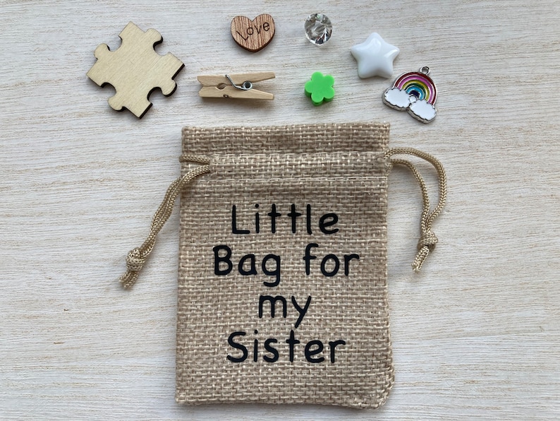 Może przedstawiać: Mały jutowy woreczek ze sznurkiem do zamykania. Woreczek jest ozdobiony napisem "Little Bag for my Sister". Woreczek jest otoczony r&oacute;żnymi małymi przedmiotami, w tym drewnianym sercem z wygrawerowanym napisem "Love", małą zieloną koniczyną, białą gwiazdą, zawieszką w kształcie tęczy i drewnianym elementem układanki.