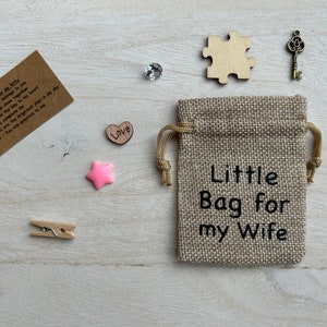 May include: A small burlap bag with the text "Little Bag for my Wife" printed on it. The bag is surrounded by various small items, including a wooden heart with the word "Love" on it, a pink star, a wooden puzzle piece, a small key, and a piece of paper with a message written on it.