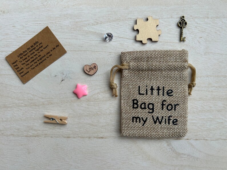 May include: A small burlap bag with the text "Little Bag for my Wife" printed on it. The bag is surrounded by various small items, including a wooden heart with the word "Love" on it, a pink star, a wooden puzzle piece, a small key, and a piece of paper with a message written on it.