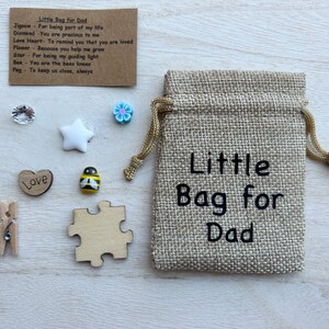 May include: A small hessian bag with the text "Little Bag for Dad" printed on it. The bag contains a variety of small items, including a wooden heart with the word "Love" on it, a wooden jigsaw piece, a small wooden star, a small bee figurine, a small blue flower, and a small clear crystal.
