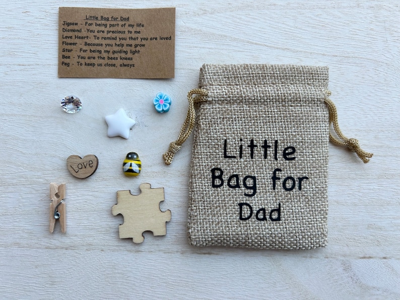 May include: A small hessian bag with the text "Little Bag for Dad" printed on it. The bag contains a variety of small items, including a wooden heart with the word "Love" on it, a wooden jigsaw piece, a small wooden star, a small bee figurine, a small blue flower, and a small clear crystal.
