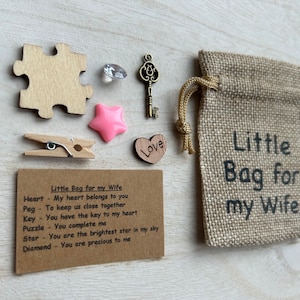 May include: A small burlap bag with the text "Little Bag for my Wife" printed on it. The bag contains a wooden heart, a wooden peg, a wooden puzzle piece, a wooden key, a pink star, and a diamond. A card with the text "Little Bag for my Wife" and a list of items and their meanings is included.