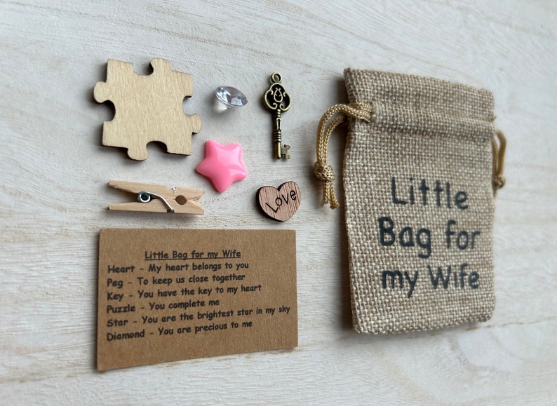 May include: A small burlap bag with the text "Little Bag for my Wife" printed on it. The bag contains a wooden heart, a wooden peg, a wooden puzzle piece, a wooden key, a pink star, and a diamond. A card with the text "Little Bag for my Wife" and a list of items and their meanings is included.