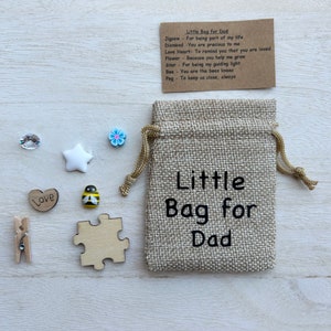 May include: A small hessian bag with the text "Little Bag for Dad" printed on it. The bag contains a variety of small items, including a wooden jigsaw piece, a wooden heart with the word "Love" on it, a wooden bee, a white star, a blue flower, a clear rhinestone, and a wooden peg.