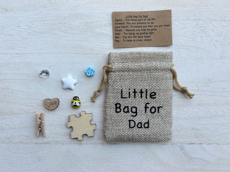 May include: A small hessian bag with the text "Little Bag for Dad" printed on it. The bag contains a variety of small items, including a wooden jigsaw piece, a wooden heart with the word "Love" on it, a wooden bee, a white star, a blue flower, a clear rhinestone, and a wooden peg.
