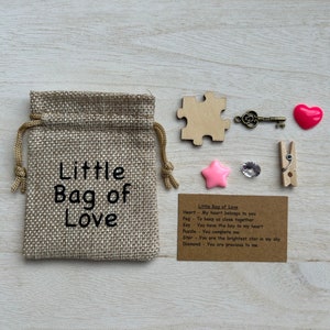 May include: A small burlap bag with the text "Little Bag of Love" printed on it. The bag is filled with small items, including a wooden heart, a key, a puzzle piece, a pink star, a clear rhinestone, and a wooden clothespin. A card with the text "Little Bag of Love" and a list of items with their meanings is also included.