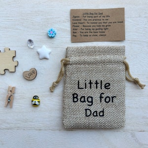 May include: A small hessian bag with the text "Little Bag for Dad" printed on it. The bag is surrounded by small decorative items, including a wooden heart with the word "Love" on it, a wooden jigsaw puzzle piece, a white star, a blue flower, a bee, a small wooden peg, and a clear crystal.