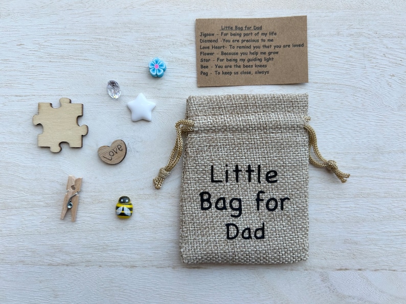 May include: A small hessian bag with the text "Little Bag for Dad" printed on it. The bag is surrounded by small decorative items, including a wooden heart with the word "Love" on it, a wooden jigsaw puzzle piece, a white star, a blue flower, a bee, a small wooden peg, and a clear crystal.