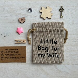 May include: A small burlap bag with the text "Little Bag for my Wife" printed on it. The bag contains a variety of small items, including a heart, a peg, a key, a puzzle piece, a star, and a diamond. The items are accompanied by a card with the text "Little Bag for my Wife" and a description of each item.
