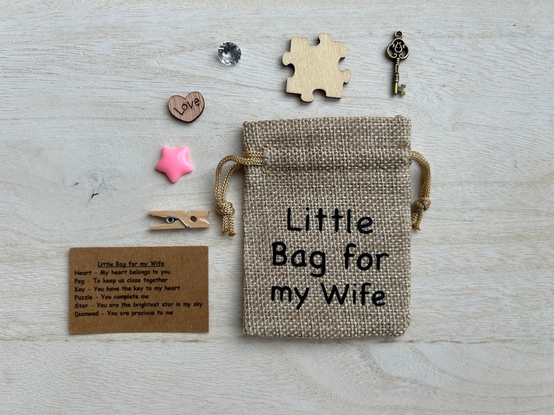 May include: A small burlap bag with the text "Little Bag for my Wife" printed on it. The bag contains a variety of small items, including a heart, a peg, a key, a puzzle piece, a star, and a diamond. The items are accompanied by a card with the text "Little Bag for my Wife" and a description of each item.