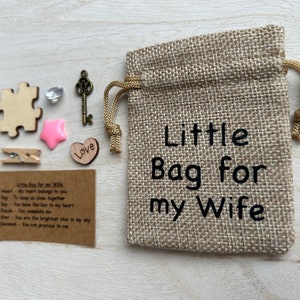 May include: A small burlap bag with the text "Little Bag for my Wife" printed on it. The bag is filled with small items, including a wooden heart, a wooden puzzle piece, a wooden star, a small key, a crystal, and a wooden clothespin. A card with the text "Little Bag for my Wife" and a list of items with their meanings is also included.
