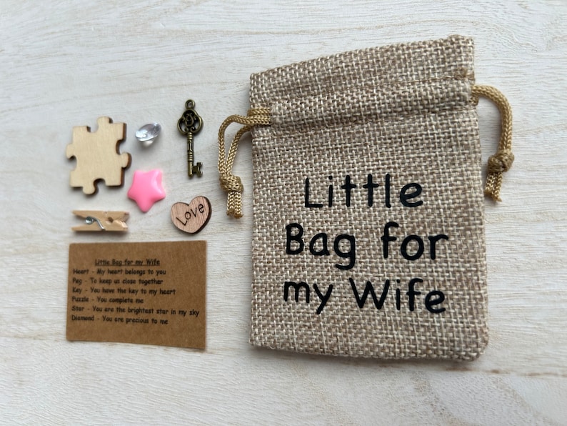 May include: A small burlap bag with the text "Little Bag for my Wife" printed on it. The bag is filled with small items, including a wooden heart, a wooden puzzle piece, a wooden star, a small key, a crystal, and a wooden clothespin. A card with the text "Little Bag for my Wife" and a list of items with their meanings is also included.
