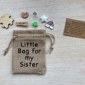 Może przedstawiać: Mały woreczek z juty z nadrukiem "Little Bag for my Sister". Woreczek jest wypełniony małymi ozdobami, w tym kawałkiem układanki, drewnianym sercem, zieloną koniczyną, gwiazdą, tęczą i kartką z tekstem "Little Bag for my Sister" oraz listą przedmiot&oacute;w z ich znaczeniami.