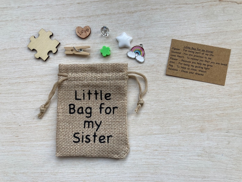 Może przedstawiać: Mały woreczek z juty z nadrukiem "Little Bag for my Sister". Woreczek jest wypełniony małymi ozdobami, w tym kawałkiem układanki, drewnianym sercem, zieloną koniczyną, gwiazdą, tęczą i kartką z tekstem "Little Bag for my Sister" oraz listą przedmiot&oacute;w z ich znaczeniami.