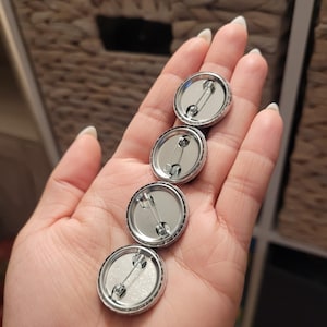 May include: Four silver-colored pinback buttons with safety pin closures are held in a hand. The buttons have a shiny, reflective surface and a simple, classic design. The background includes a woven basket and a white shelf.