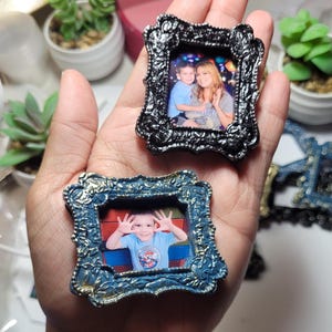 May include: Two miniature ornate picture frames, one black and one blue, each holding a small photograph. The black frame has a detailed floral design. The blue frame has a gold trim. The frames are held in a hand.