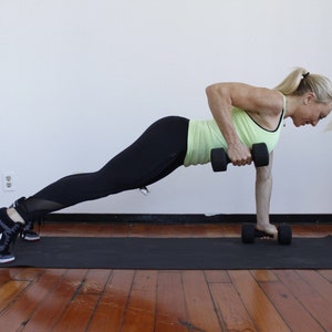 May include: A woman in a green tank top and black leggings performs a dumbbell row exercise while in a plank position. She is using a black dumbbell in each hand.