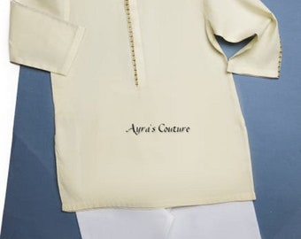 Puede incluir: Una camisa de manga larga de color crema con cuello mandarín y ribetes dorados. La camisa se combina con pantalones blancos. El texto "Ayra's Couture" está impreso en la camisa.