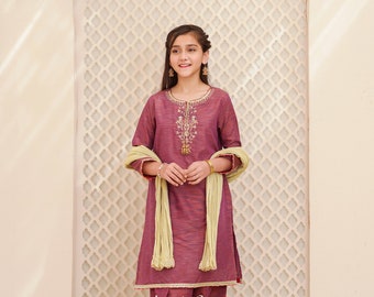 Puede incluir: Una persona joven lleva un salwar kameez granate con bordados dorados y un dupatta verde claro. El atuendo se combina con pantalones granates y ribetes dorados. La persona está de pie frente a una pared de celosía blanca.