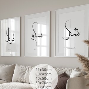 May include: Three white framed prints with black calligraphy in Arabic script. The prints are hung on a white wall above a beige couch. The text in the prints reads "Thank you", "Peace", and "Grateful".