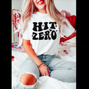 May include: A white short-sleeved t-shirt featuring the black text HIT ZERO in a bold, wavy, retro-inspired font. The design includes small star accents and a heart shape integrated into the letter O. The shirt is paired with light blue denim jeans.