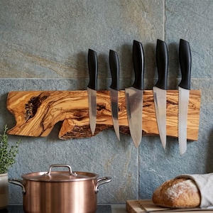 May include: A wooden knife holder with six knives. The holder is made of a light brown wood with a unique grain pattern. The knives have black handles and silver blades. The holder is mounted on a gray tiled wall. A copper pot and bread are in the foreground.