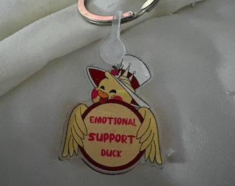 May include: A keychain with a cartoon duck wearing a chef's hat and holding a sign that reads "EMOTIONAL SUPPORT DUCK". The duck has yellow wings and a red and white sign. The keychain has a silver key ring.