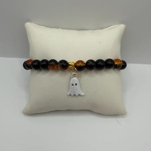 May include: A beaded bracelet featuring alternating black and amber-coloured round beads with a central gold-toned spacer bead. A small, white, enamel-style ghost charm hangs from the centre of the bracelet, attached by a gold-toned jump ring.