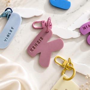 May include: A collection of colourful letter-shaped keychains featuring names such as VIOLET, KENZIE, ELI, MIA, and EMMA. Each keychain includes a matching metal lobster clasp and is paired with a white feather-shaped charm, showcasing various pastel and bold hues.