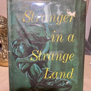 Könnte beinhalten: Ein Vintage-Hardcover-Buch mit dem Titel "Stranger in a Strange Land" von Robert A. Heinlein. Der Umschlag zeigt einen grünen Hintergrund mit einer bronzenen Skulptur einer Person, die mit dem Kopf in den Händen sitzt.