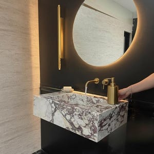 May include: A modern bathroom sink made of marble with a purple and white pattern. The sink has a gold faucet and soap dispenser. A round mirror with a light is above the sink.