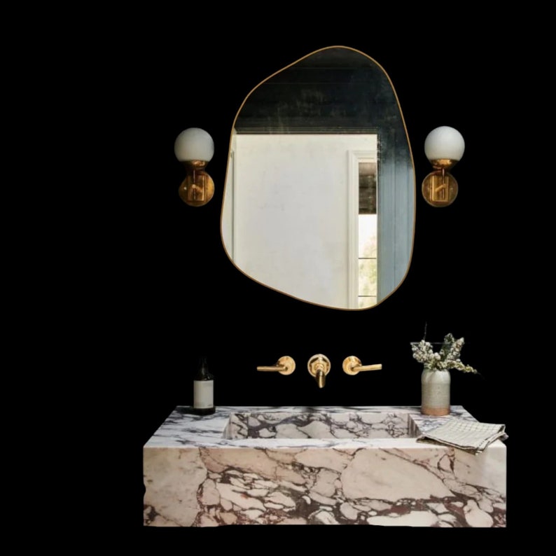 A calacatta viola marble bathroom vanity  single sink with a gold faucet and two gold wall sconces with white glass shades. The vanity has a white marble countertop and a white marble sink. A small plant is in a white vase on the countertop. The mirror above the sink is a gold oval frame with a unique shape. The background is a dark black wall.