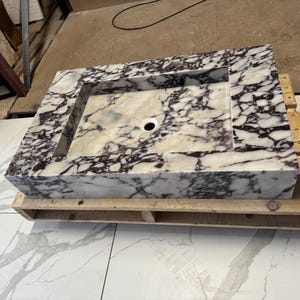 A calacatta viola marble sink with brown and purple veins. The sink is rectangular and has a square basin. The sink is sitting on a white surface.