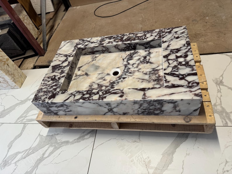 A calacatta viola marble sink with brown and purple veins. The sink is rectangular and has a square basin. The sink is sitting on a white surface.