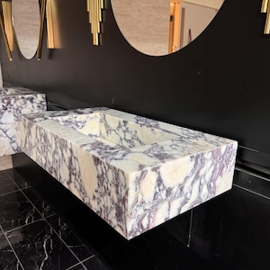 A calacatta viola marble sink with brown and purple veins. The sink is rectangular and has a square basin. The sink is sitting on a white surface.