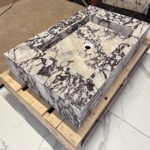 A calacatta viola marble sink with brown and purple veins. The sink is rectangular and has a square basin. The sink is sitting on a white surface.