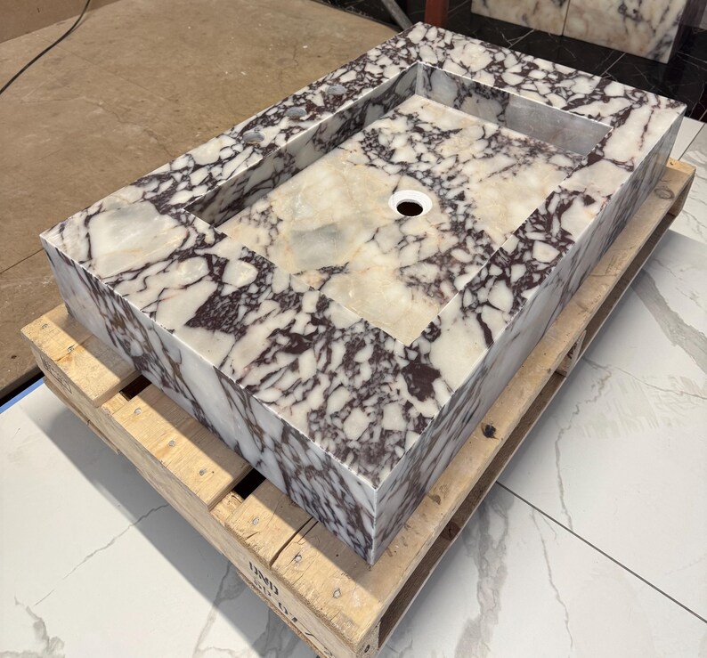 A calacatta viola marble sink with brown and purple veins. The sink is rectangular and has a square basin. The sink is sitting on a white surface.