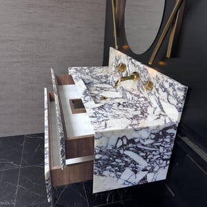 A white and purple marble bathroom vanity with two drawers. The vanity has a single sink and gold faucets. The vanity is mounted on a black tiled floor.