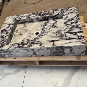A calacatta viola marble sink with brown and purple veins. The sink is rectangular and has a square basin. The sink is sitting on a white surface.