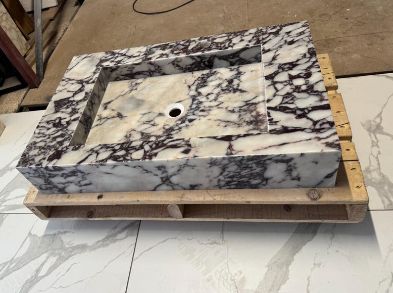 A calacatta viola marble sink with brown and purple veins. The sink is rectangular and has a square basin. The sink is sitting on a white surface.