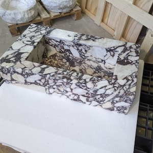 A calacatta viola marble sink with brown and purple veins. The sink is rectangular and has a square basin. The sink is sitting on a white surface.