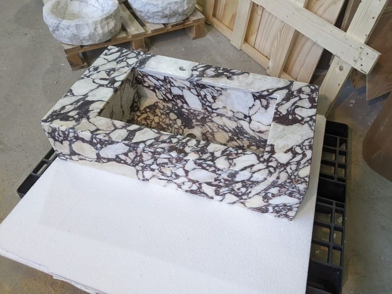 A calacatta viola marble sink with brown and purple veins. The sink is rectangular and has a square basin. The sink is sitting on a white surface.