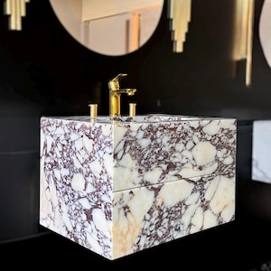 May include: A square marble sink with a gold faucet and fixtures. The sink features a white and burgundy patterned design. The faucet and fixtures are gold. The sink is set against a black wall with decorative gold accents.