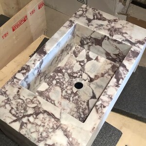 A calacatta viola marble sink with a rectangular shape and a drain hole in the center. The marble has a light gray background with dark gray and purple veins throughout.
