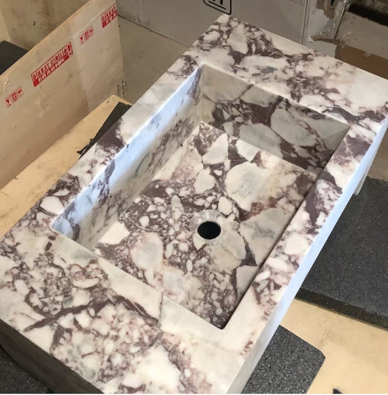 A calacatta viola marble sink with a rectangular shape and a drain hole in the center. The marble has a light gray background with dark gray and purple veins throughout.