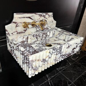 A calacatta viola marble sink with brown and purple veins. The sink is rectangular and has a square basin. The sink is sitting on a white surface.